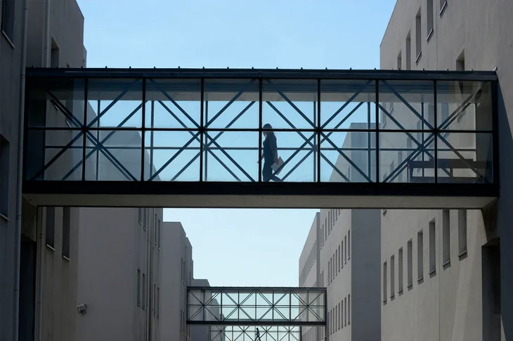Uma mulher caminha numa ponte transparente, entre dois edifícios. Mais pontes iguais surgem em perspetiva no fundo da imagem.