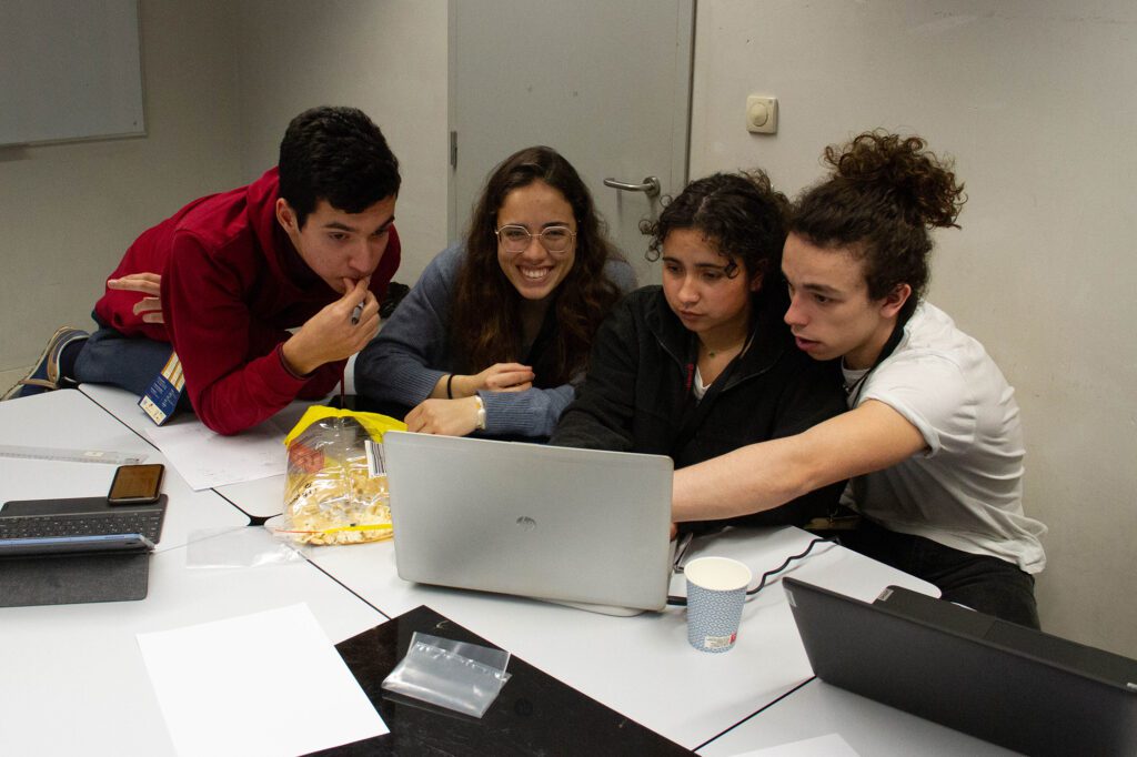 4 estudantes a olhar para um computador com ar entusiasmado. Papéis e um protótipo pela mesa.
