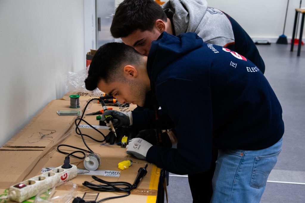 Estudantes de engenharia debruçados sobre uma mesa com fios elétricos, a observar um protótipo.