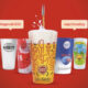 Um copo de cerveja dourada Super Bock sendo servido, com espuma no topo e design gráfico vibrante, destacado contra um fundo vermelho, cercado por outros copos temáticos de festivais de música patrocinados pela Super Bock.