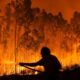 Morador tentando combater um dos focos de incêndio florestal durante a noite, em uma área densamente arborizada, envolta por chamas intensas. A silhueta da pessoa contrasta com o brilho alaranjado do fogo.