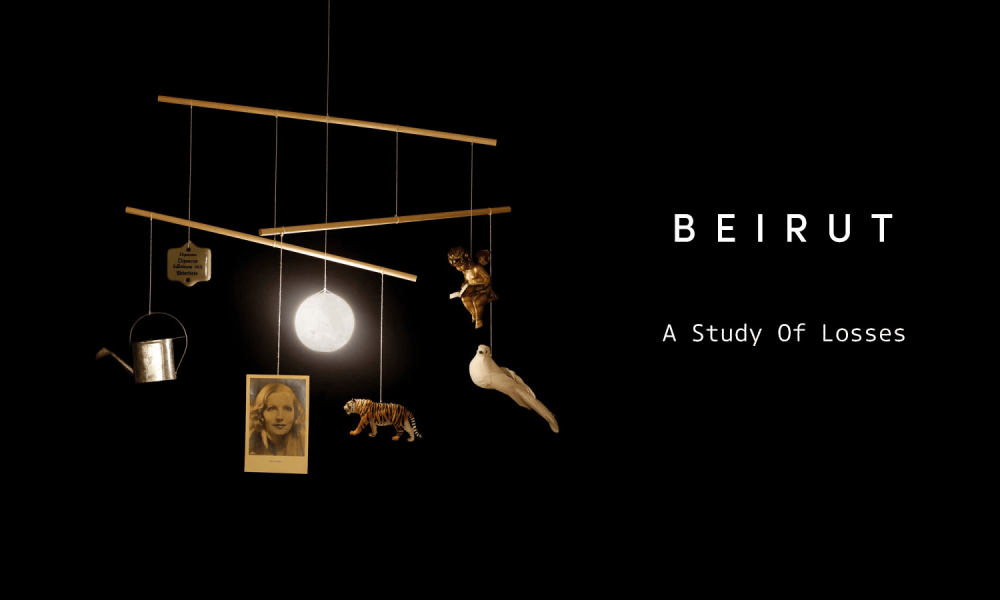 Capa do álbum A Study of Losses, da banda Beirut, mostrando um móbile suspenso sobre fundo preto. Os objetos pendurados — uma pequena lata de regador metálico, uma fotografia antiga de uma mulher, um tigre em miniatura, um anjo dourado, uma pomba branca e uma esfera luminosa — criam um equilíbrio delicado e simbólico. À direita, o nome “Beirut” e o título do álbum aparecem em tipografia branca minimalista.