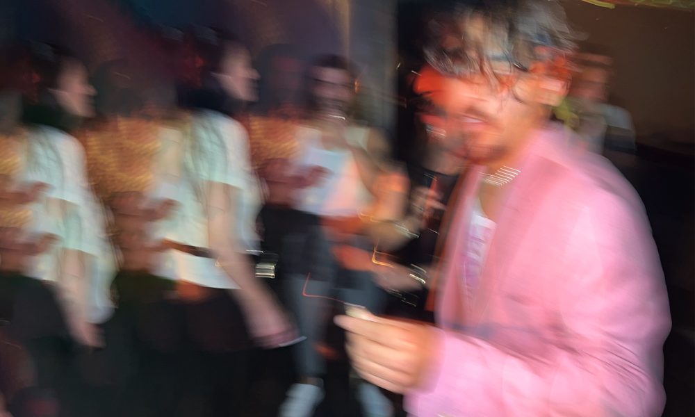 Imagem desfocada de uma festa ou bar, com várias pessoas ao fundo em movimento. Em primeiro plano, há uma pessoa usando um blazer rosa, também desfocada, segurando algo na mão. As luzes coloridas criam um efeito de longa exposição, deixando rastros e duplicações das figuras. Comemorasse os meus 32 anos de vida.