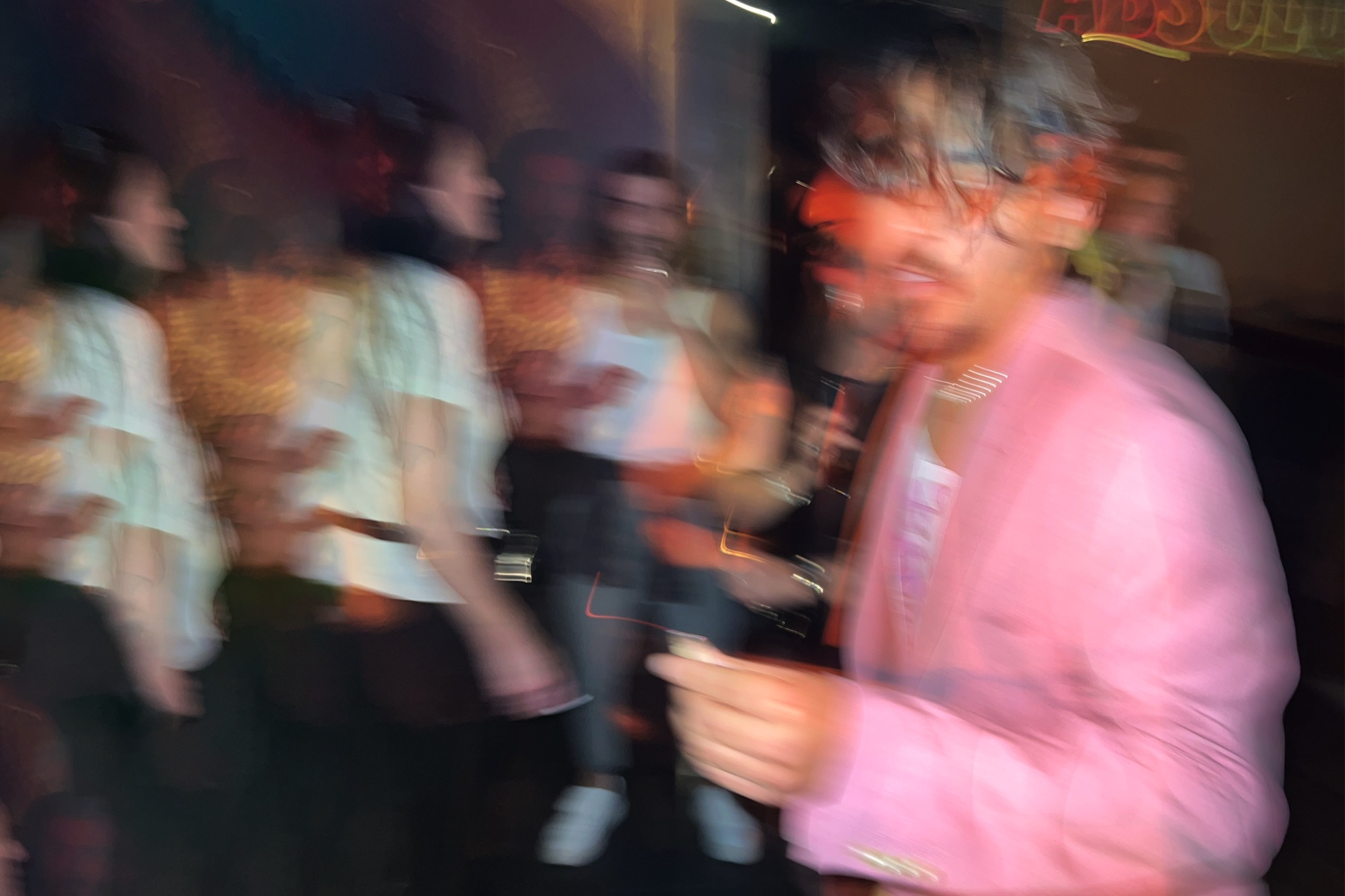 Imagem desfocada de uma festa ou bar, com várias pessoas ao fundo em movimento. Em primeiro plano, há uma pessoa usando um blazer rosa, também desfocada, segurando algo na mão. As luzes coloridas criam um efeito de longa exposição, deixando rastros e duplicações das figuras. Comemorasse os meus 32 anos de vida.